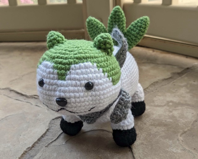 Crocheted Sommie in aromantic flag colours: green face mask and tail, black paws, and gray scarf.